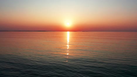 Vista-Aérea-Por-Drones-Del-Hermoso-Amanecer-En-El-Océano-Con-Un-Sol-Brillante-Que-Proyecta-Coloridos-Reflejos-Rojos,-Naranjas,-Morados-Y-Amarillos-Sobre-Las-Tranquilas-Olas-Del-Mar-Con-Un-Hermoso-Cielo-Y-Agua