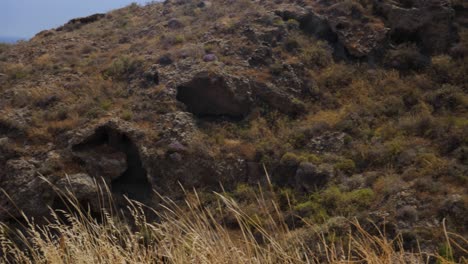 Toma-Panorámica-De-Mano-De-Unas-Cuantas-Cuevas-Misteriosas-En-Una-Colina-De-Montaña-Cerca-De-La-Playa-Roja-En-Santorini,-Grecia