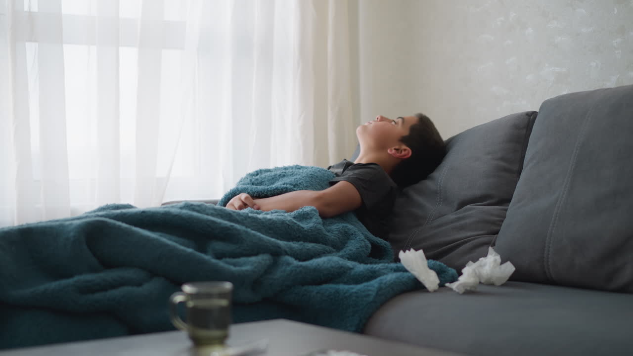 Un bambino piccolo che dorme su un divano avvolto in una coperta, si sistema per stare più comodo con dei fazzoletti sul divano; un momento accogliente e tranquillo in casa con luce naturale dalla finestra.