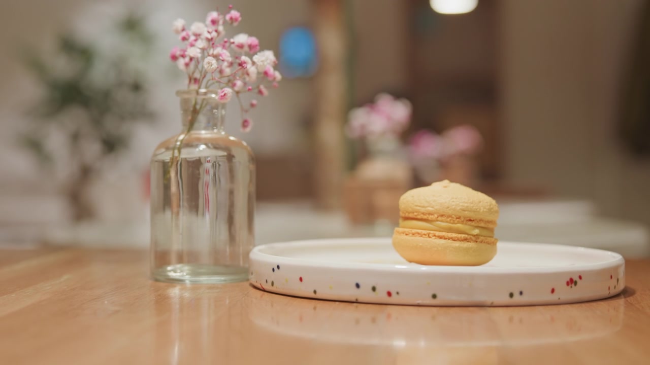 Close up of hand reaching towards and gently touching empty glass vase with pink flowers then moving away. macaron is placed on white plate beside vase by another person hands in soft indoor