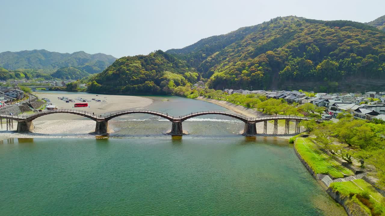 A beautiful, wide aerial panning shot showcasing the full length of the iconic Kintai Bridge in Iwakuni, Japan. The video captures the five wooden arches spanning the clear Nishiki River,