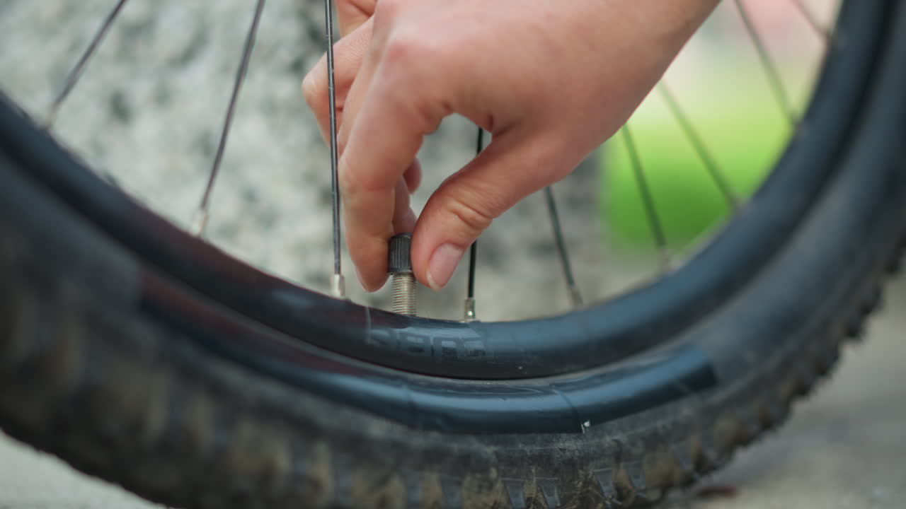woman repairing bicycle valve on muddy tire, closeup of fingers unscrewing cap and checking rim grimy spokes, concrete sidewalk, patient roadside fix showing tension and relief in precise motion