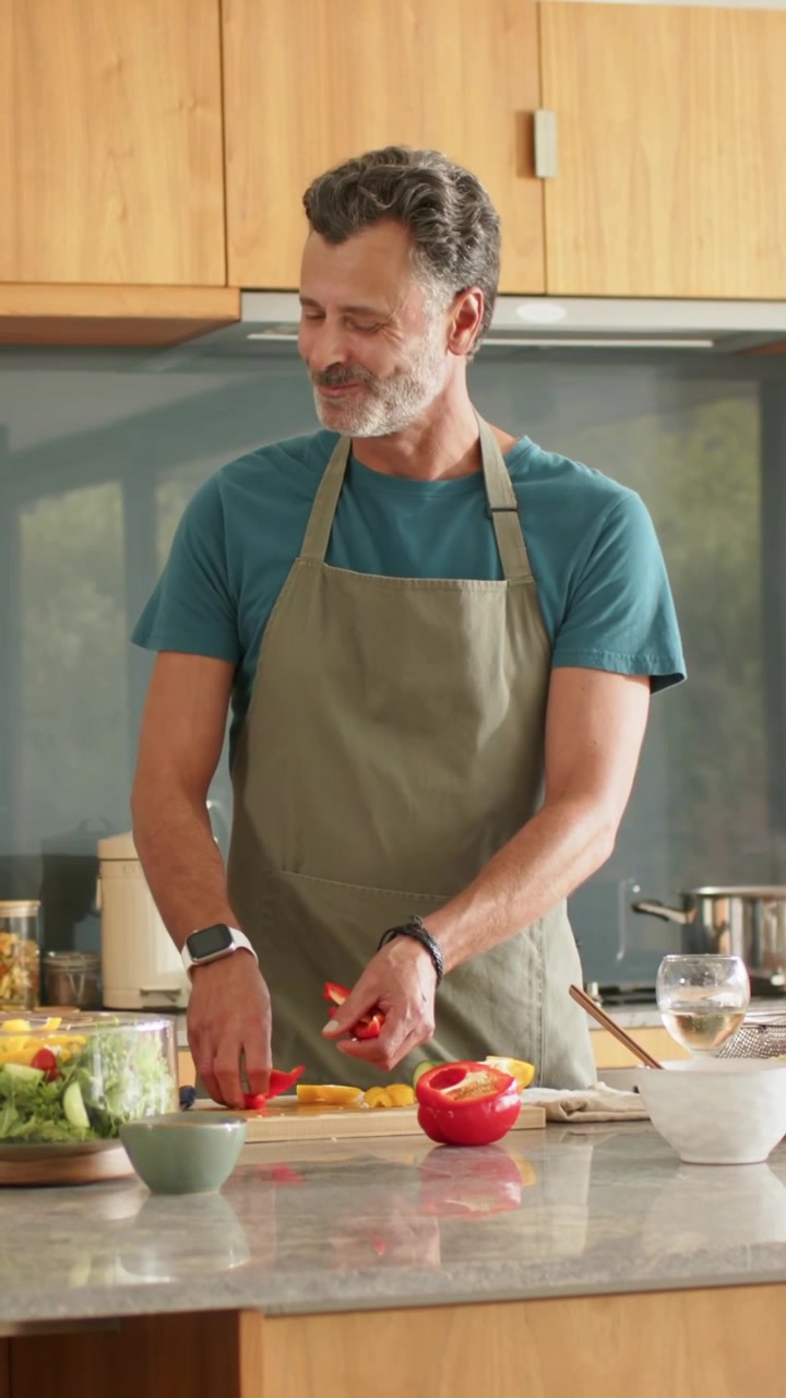 Vertikales Video: Mann in Schürze schneidet Paprika an der Kücheninsel für Salat, während der Partner eintrifft