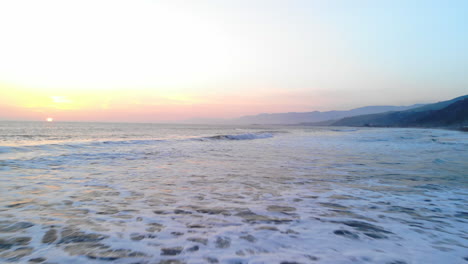 Toma-Aérea-Volando-Bajo-Sobre-Las-Olas-Del-Océano-Rompiendo-En-La-Playa-Bajo-Un-Suave-Fondo-De-Cielo-De-Puesta-De-Sol-En-La-Costa-De-Ventura,-Drone-De-California