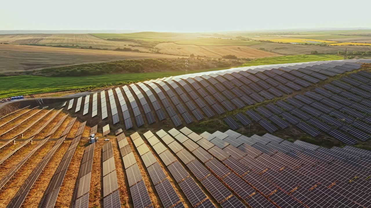 Solpaneler opsamler energi på en mark ved solnedgang nær et landområde med åbne landskaber i baggrunden.