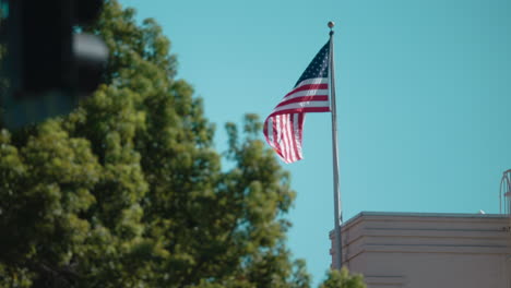 Eine-Aufnahme-Einer-Amerikanischen-Flagge,-Die-Im-Wind-Auf-Dem-Dach-Eines-Gebäudes-Flattert,-Mit-Einem-Baum-Und-Einer-Ampel-Im-Vordergrund-Bei-Tag