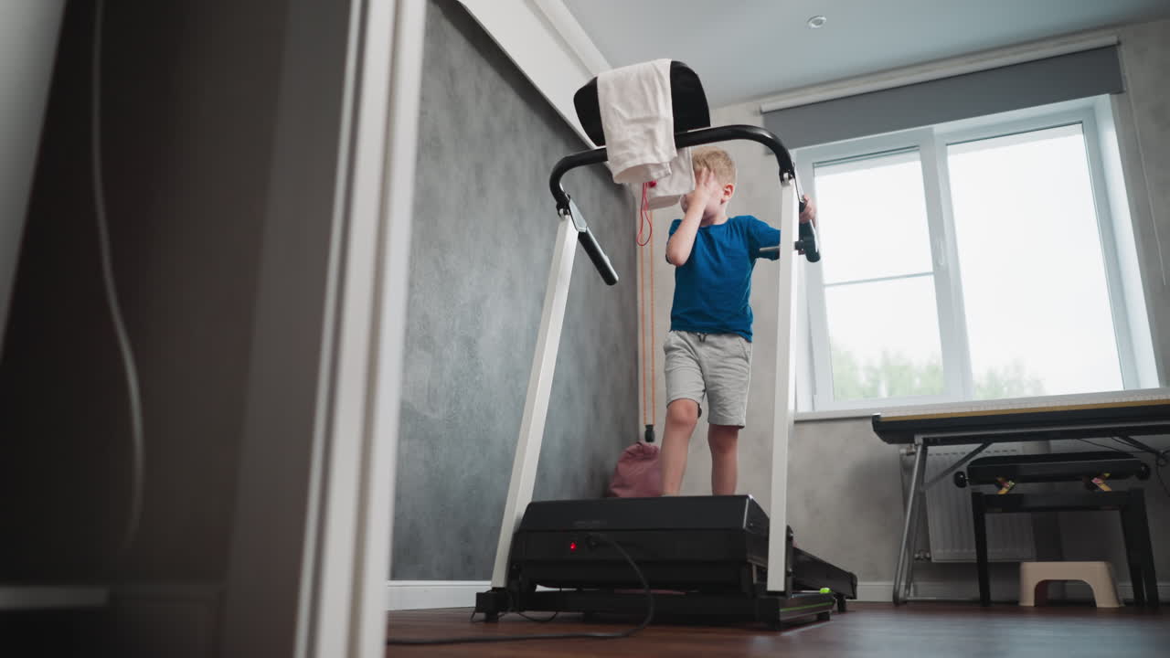 little boy exercising on treadmill gripping handle while cleaning face in brightly lit home gym environment with window and keyboard in background reflecting natural indoor fitness activity