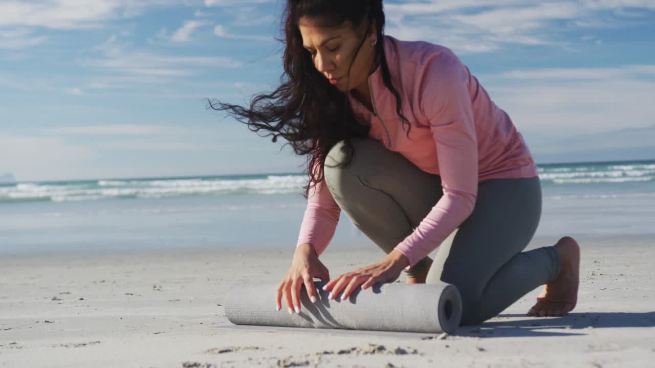 mujer de raza mixta rodando colchoneta de yoga en la playa