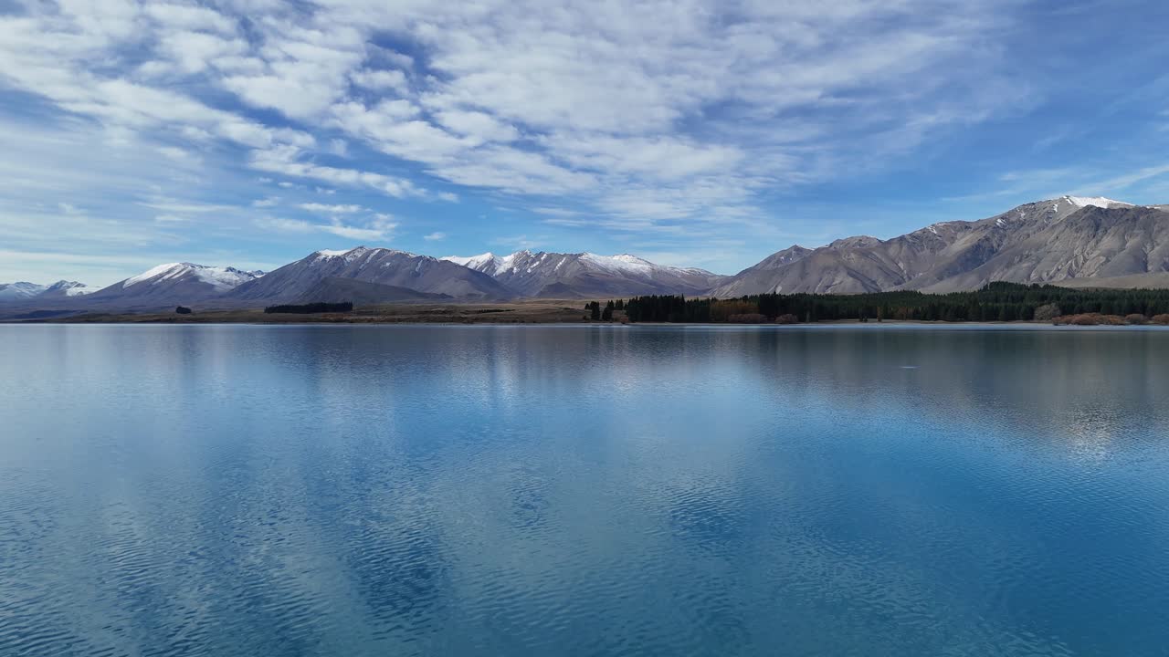Drone captures Lake Tekapo's calm waters and surrounding mountains under a vibrant sky, showcasing natural beauty and tranquility
