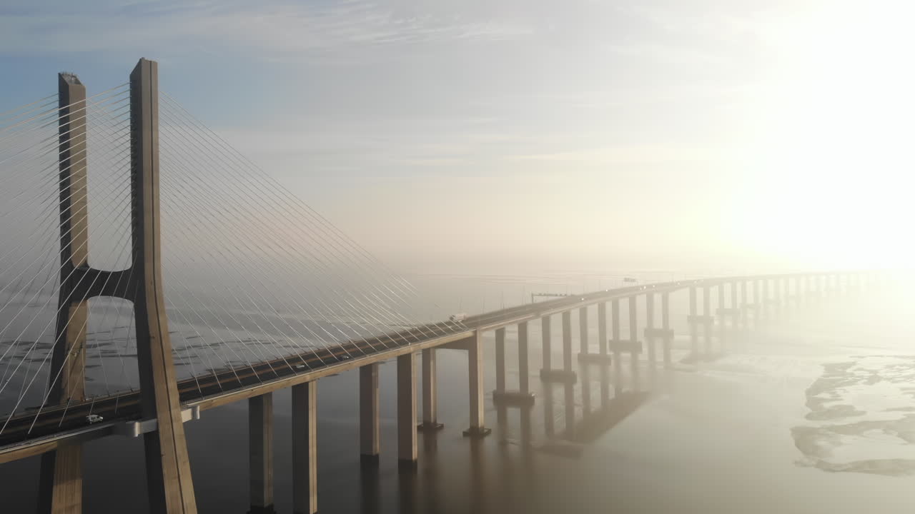 The video showcases a majestic cable-stayed bridge standing tall over tranquil water in the early morning light. Soft mist surrounds the area, creating a serene atmosphere
