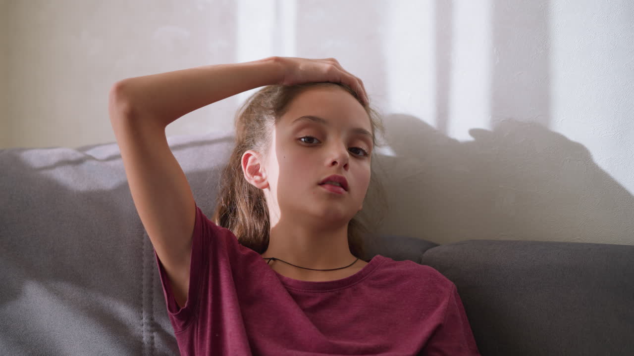 Thoughtful girl lying down on couch, hand on head, looking sick and unwell, appearing to be lost in thought while dealing with cold or flu symptoms, showing signs of discomfort and fatigue