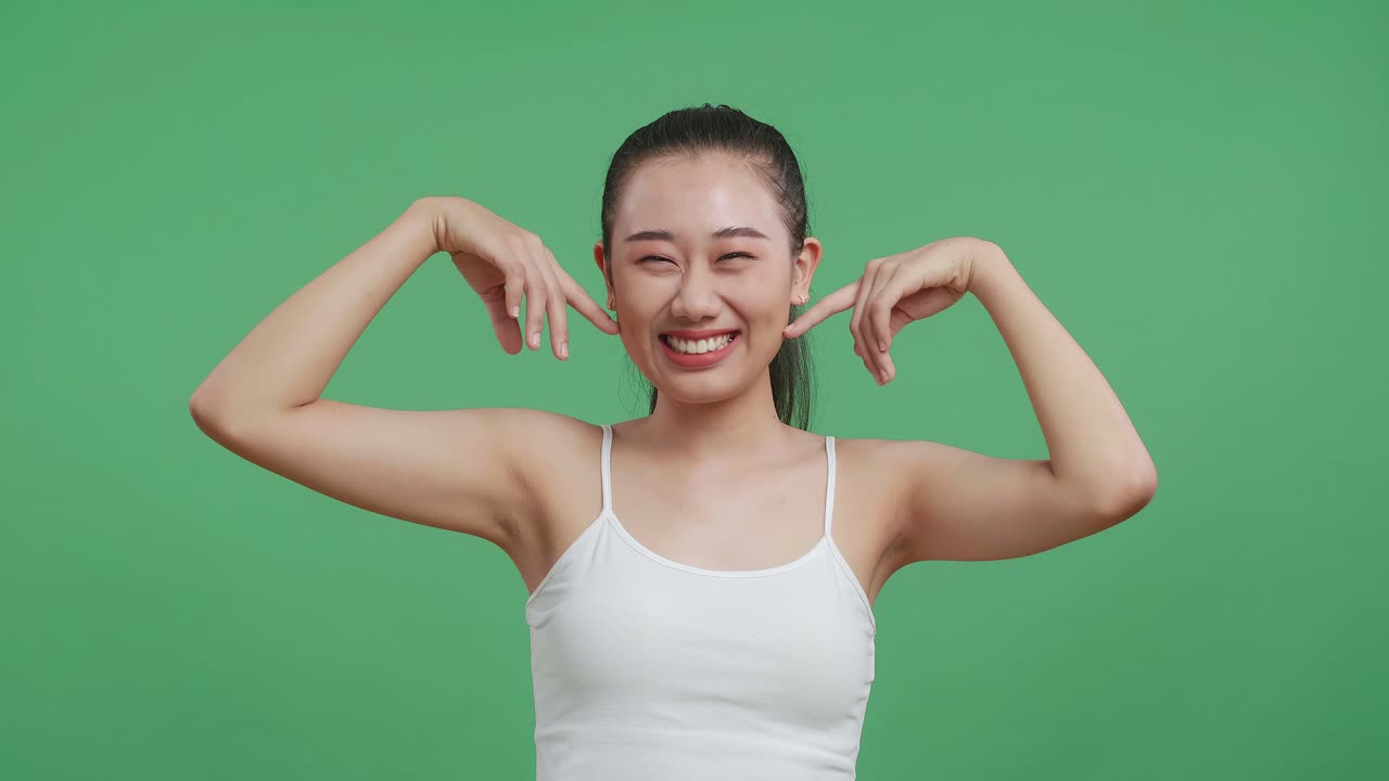 색 카미솔을 입은 아시아 여성이 인스 손가락으로 얼굴을 만지고 스튜디오의 녹색 스크린 배경에 미소 짓고 있습니다.