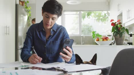 caucasian woman taking notes and calculating finances using smartphone at home