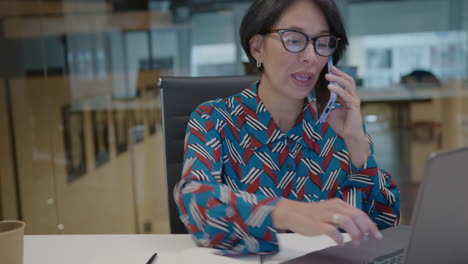 mature businesswoman using laptop and speaking on phone in office
