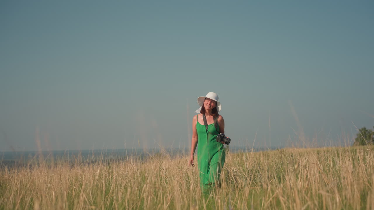 Confident stylish woman in white sunhat and green sundress walking alone through tall golden grass under clear blue sky while holding camera, embodying calm strength