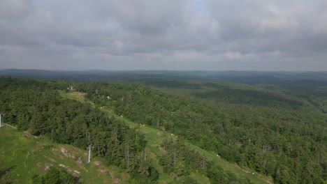 aerial shot flying over thick green tree covered ski mountain with chairlift on a cloudy summer day. calabogie ontario