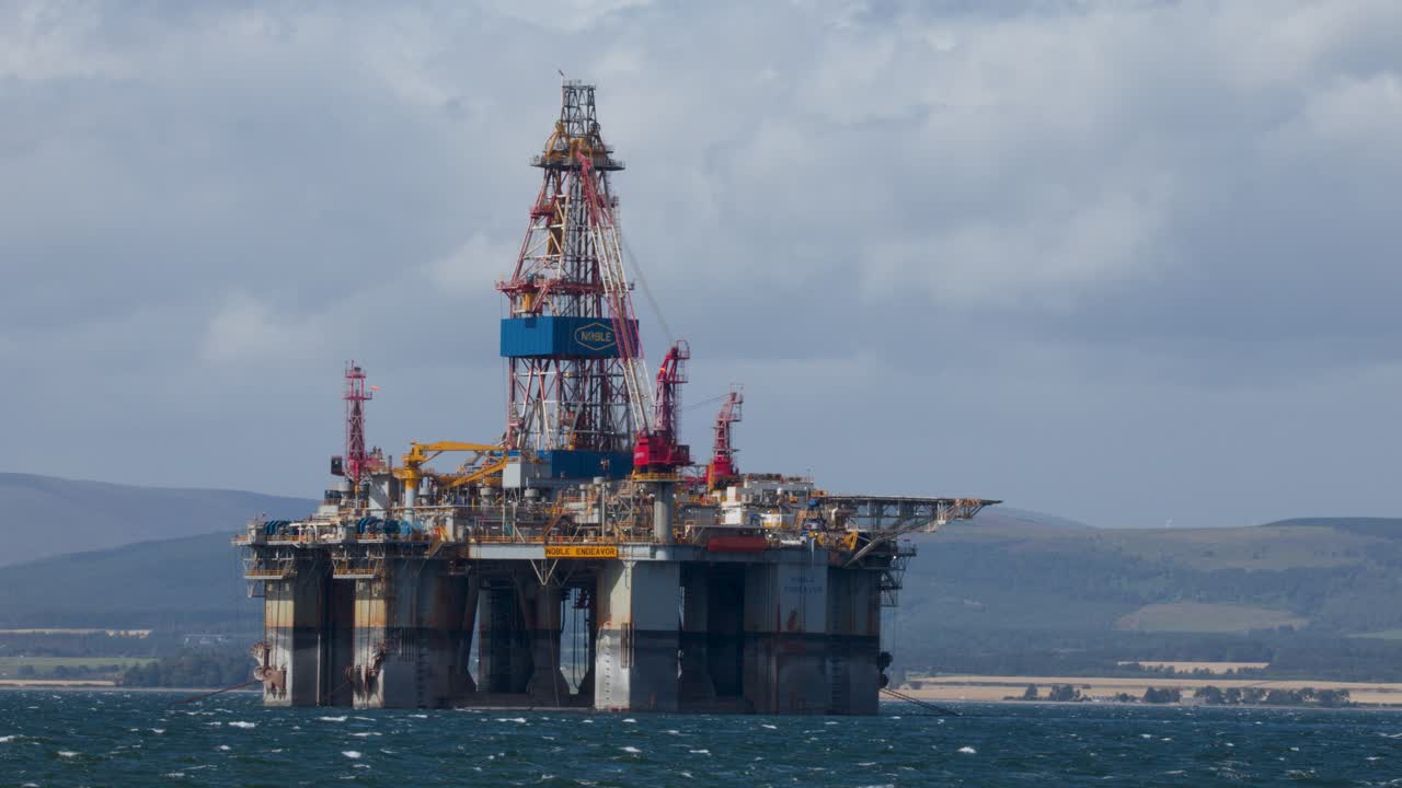 Stor offshore olieplatform stationær i oprørt hav, overskyet dagslys, fjerne bakker, stabilt kamera
