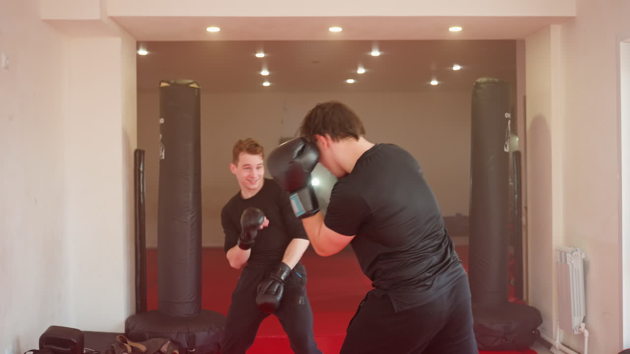 MMA fighter training inside gym facing opponent in sparring stance wearing black gloves and sportswear, preparing for strike on red mat with boxing bags in background under bright ceiling lights