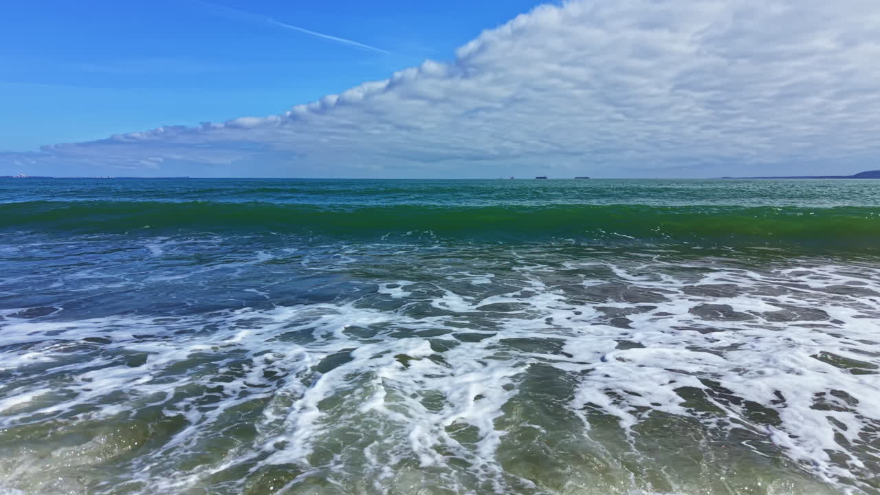 Bølger slår mot land på en åpen strand midt på dagen i et kystområde under en klar blå himmel med skyer i det fjerne
