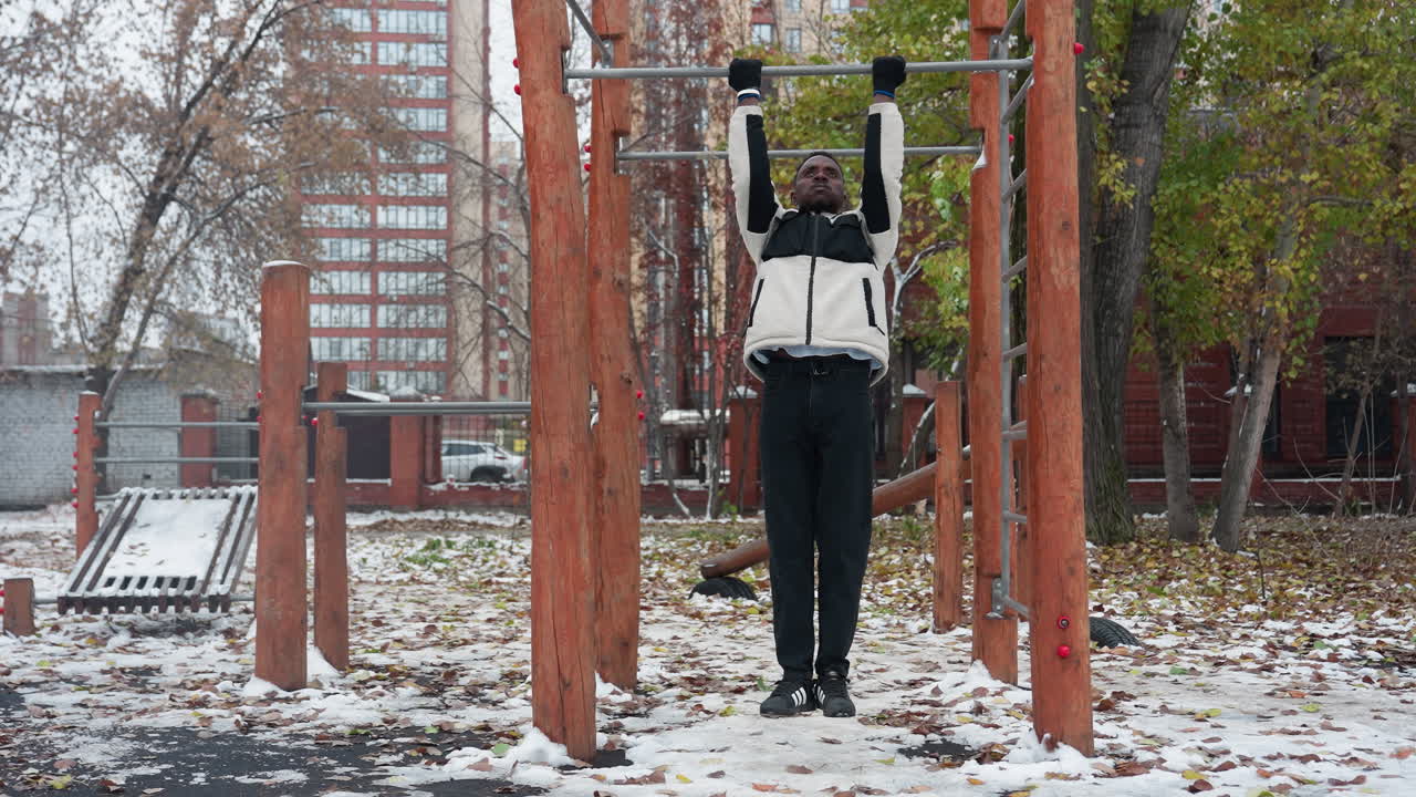 hombre realizando ejercicios de tracción en una barra de entrenamiento al aire libre en clima frío, vestido con chaqueta de invierno y guantes, levanta su cuerpo con esfuerzo, suelo cubierto de nieve, hojas caídas y fondo urbano