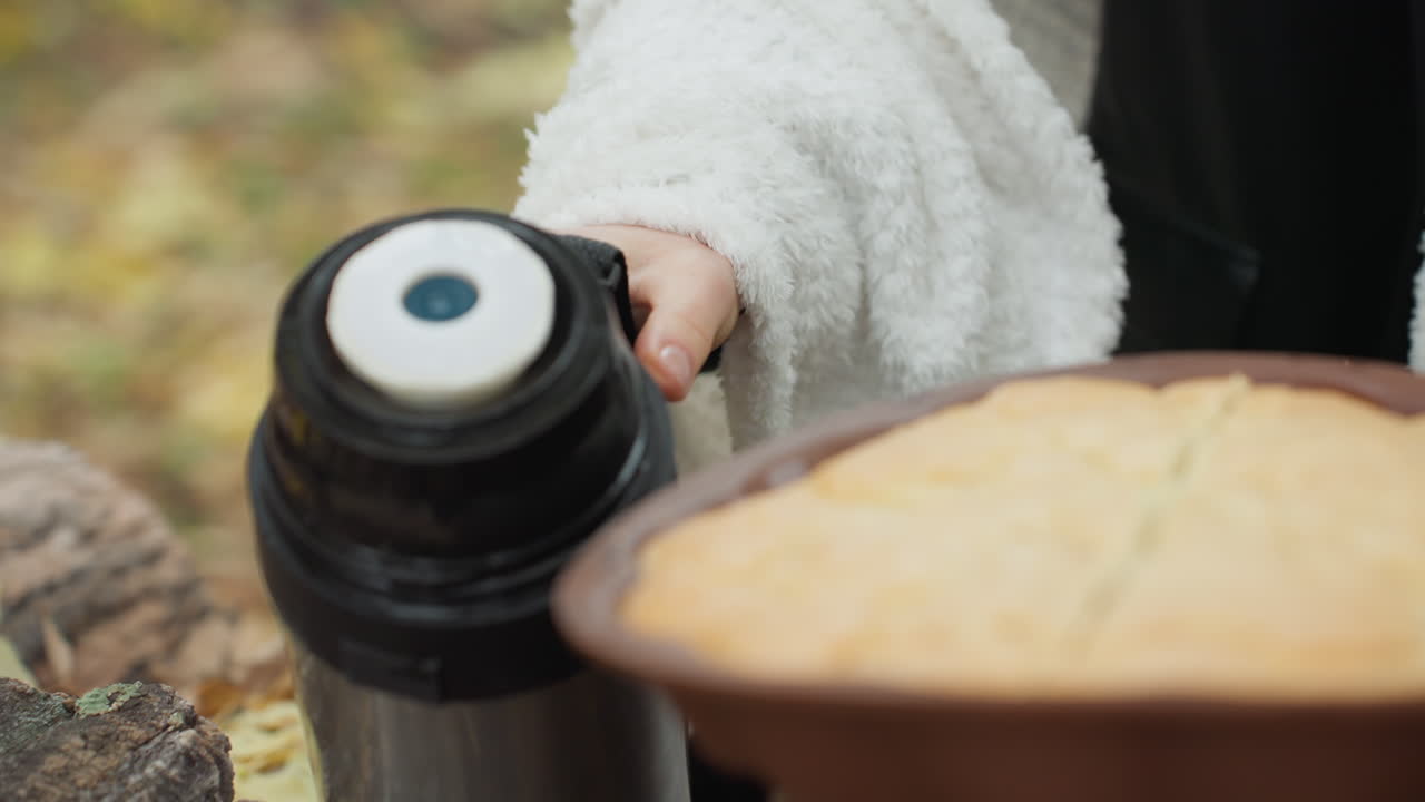 woman pouring coffee from thermos outdoors, cozy autumn picnic scene with warm mug and pie on rustic table, soft sweater draped over shoulders, closeup of hands pressing thermos lid, steam rising