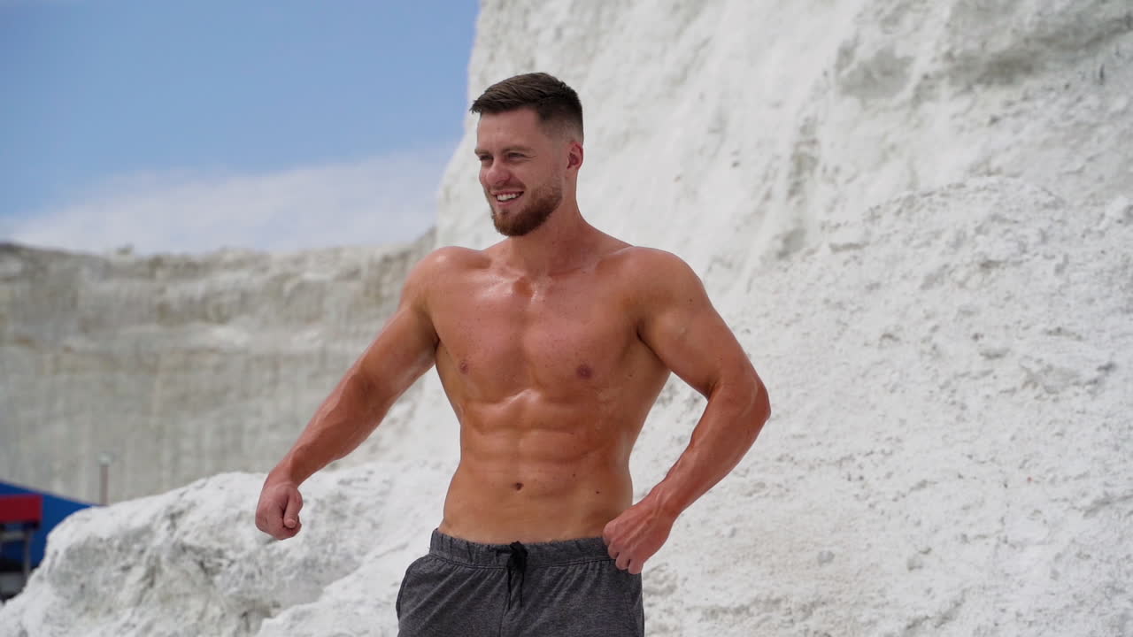 Muscular bodybuilder smiling outdoors. Strong athlete shows his trained body while standing on the white hill.
