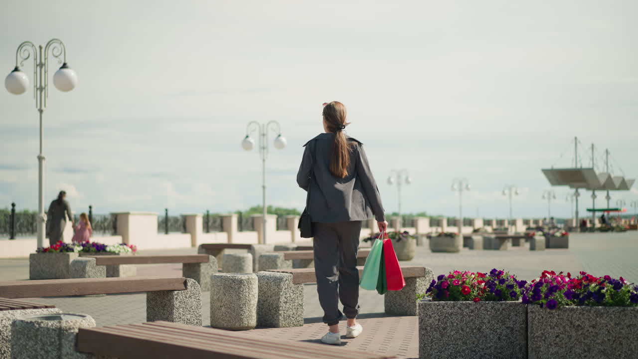 회색 옷을 입은 젊은 여자가 야외에 앉아 쇼핑 가방을 떨어뜨리고, 배경에는 어머니와 딸이 걸어가면서 손을 잡고, 멀리 있는 사람들의 흐릿한 시야가 있습니다.