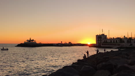 Silhouettes-of-fishermen-on-pier-catching-fish-during-orange-sunset-casting-fishing-line-on-open-sea-in-background-of-bay-and-part-of-city-in-slow-motion-capture-at-120fps