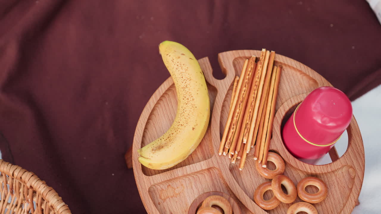 Primer plano de una mano cogiendo un plátano de una mesa de pícnic de madera llena de palitos de pan, bagels y un termo rojo sobre un mantel marrón durante un pícnic familiar al aire libre rodeado de naturaleza verde