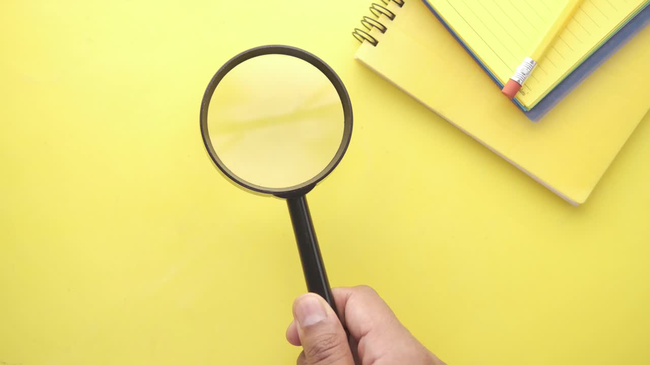 Person holding magnifying glass over yellow desk with notebook and pencil