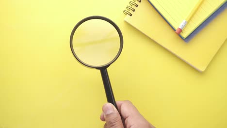 person holding magnifying glass over yellow desk with notebook and pencil