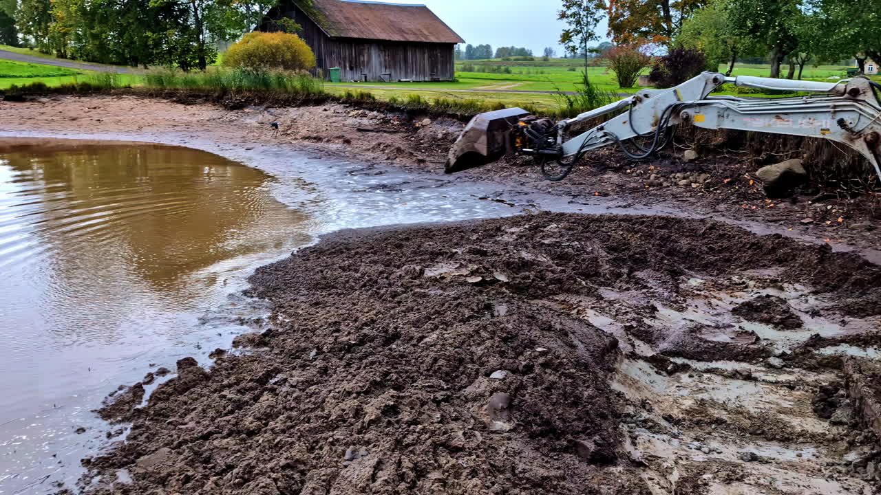 Excavator digging in muddy terrain near a grassy field on a cloudy day