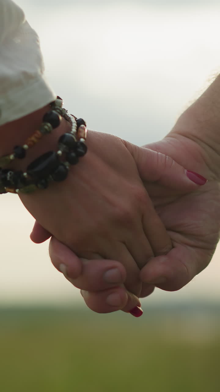 un primer plano captura las manos de dos amantes que se sostienen entre sí, con la mano de la mujer adornada con un brazalete de cuentas y uñas rojas. el sol brilla suavemente en el fondo