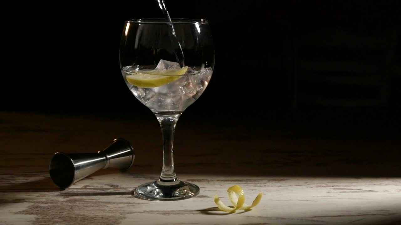 Persona che riempie un bicchiere trasparente di gin tonic con limone e jigger su un tavolo in una stanza buia
