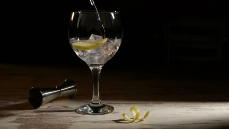 persona llenando un vaso transparente de gin tonic con limón y jigger en la mesa en una habitación oscura