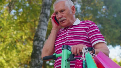 anciano anciano abuelo hombre en scooter eléctrico con bolsas de compras de colores hablando en teléfono móvil