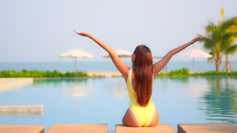 A-woman-with-her-back-to-the-camera-sits-along-the-side-of-a-swimming-pool-raising-her-arms-to-the-heavens-in-joy