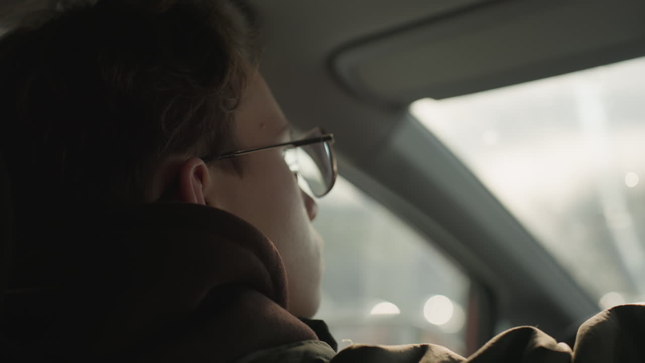 close up of young man wearing glasses inside car looking forward with soft light on face, adjusting something in interior, window reflection and bokeh light effects in background