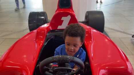 joyful scene: a 3-year-old expressive black toddler riding a red f1 simulator inside a mall