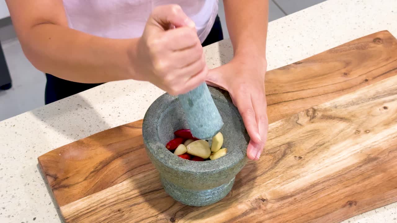 Person uses a stone mortar and pestle to crush garlic and chili on a wooden cutting board under bright, even kitchen lighting with overhead camera angle