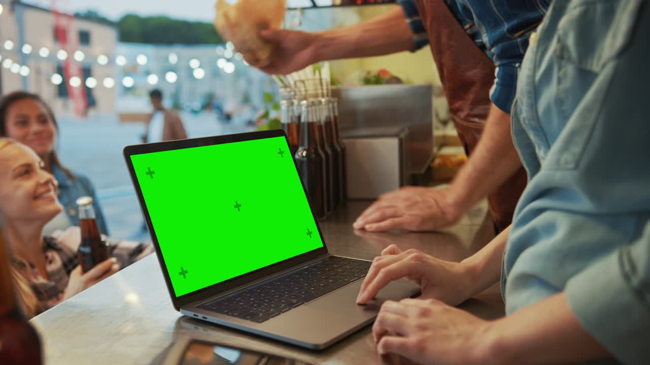 la computadora portátil con pantalla verde se coloca en un mostrador en un camión de comida callejera o quiosco que vende hamburguesas y bebidas. los clientes felices reciben sus pedidos.