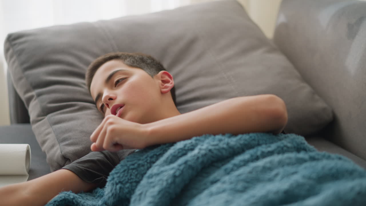 Close-up of child covered with duvet coughing, with tissue near head and light streaming through the window, the child is resting, feeling unwell