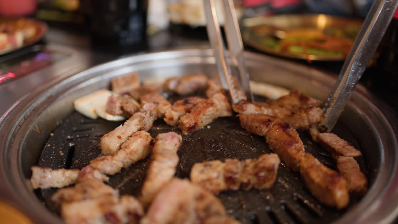 Des pinces en métal retournent et disposent des tranches de poitrine de porc assaisonnées sur un gril de table circulaire chaud.