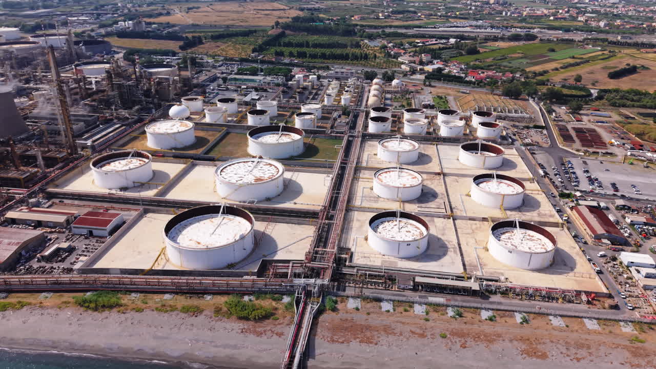 Aerial orbiting shot of oil storage tanks at a Sicilian refinery, showing pipelines, roads, and fields around the facility