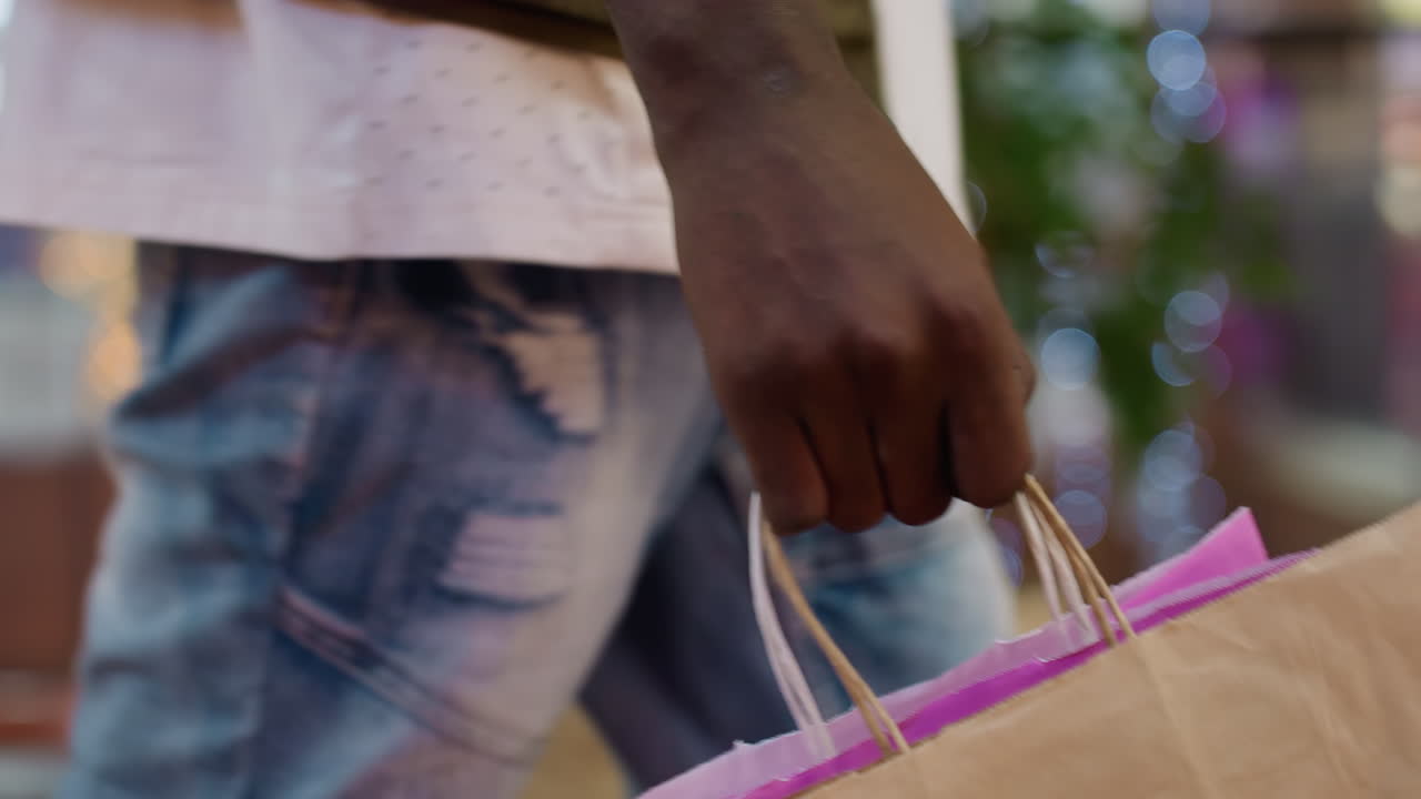 Side view of person holding paper shopping bag with firm grip while walking inside brightly lit shopping mall with blurred lights and colorful background