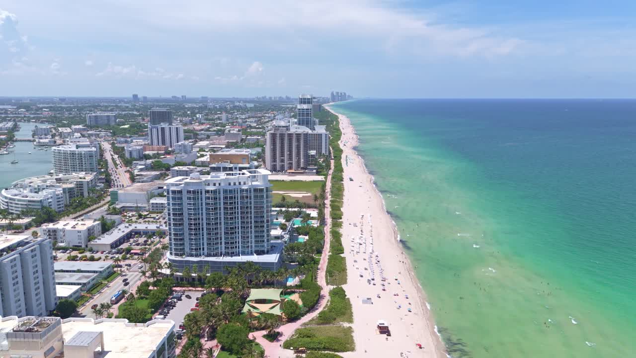 Aerial passing by high residential apartment tower with beach and ocean view Of Miami, Florida. Sunny summer day with relaxing people. Wide shot