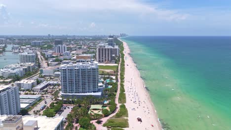 aerial passing by high residential apartment tower with beach and ocean view of miami, florida. sunny summer day with relaxing people. wide shot
