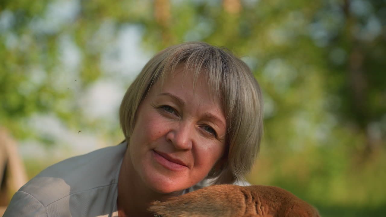 배경에 흐릿한 초록색과 밝은 하늘을 가진 갈색 개 에서 애정으로 미소 짓는 여성의 클로즈업, 야외에서 즐거운 순간을 포착