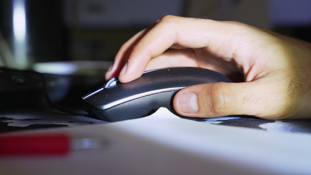 a person's hand using a computer mouse at a desk during the night. Low light creates a cinematic workspace atmosphere with blurred office supplies in the foreground