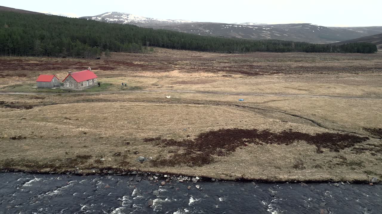 un avión no tripulado flota sobre un río frente a una plantación de pinos en las montañas de escocia y un edificio remoto con un techo rojo al lado de la pista 4x4